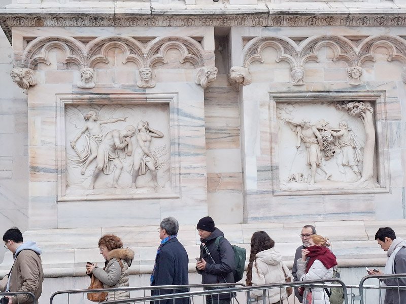 Line of visitors waiting to enter Duomo di Milano, with intricate stone carvings on the cathedral walls in the background.