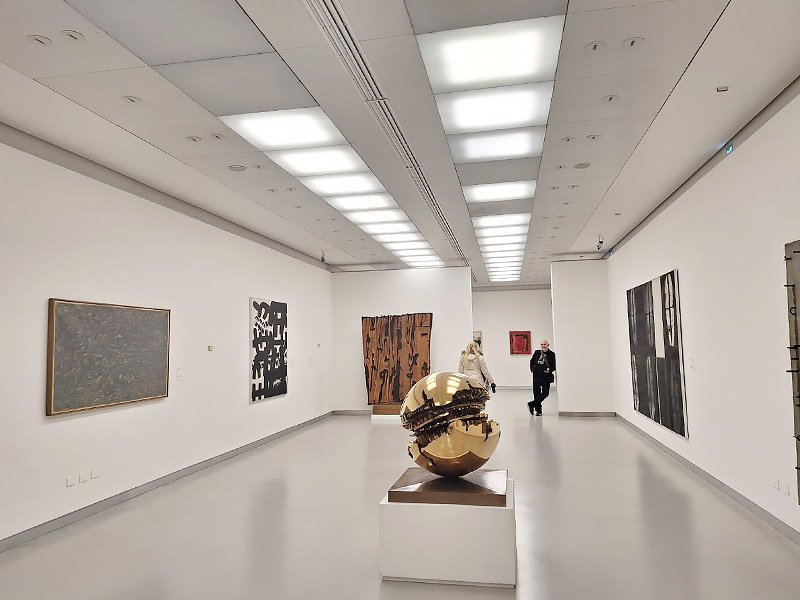 Gallery room inside Museo del Novecento in Milan featuring a large golden sphere sculpture as the centerpiece.