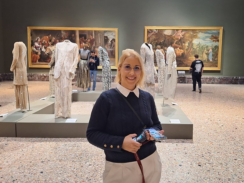 Traveler posing inside Pinacoteca di Brera in Milan, with Giorgio Armani creations and classical paintings displayed in the background.