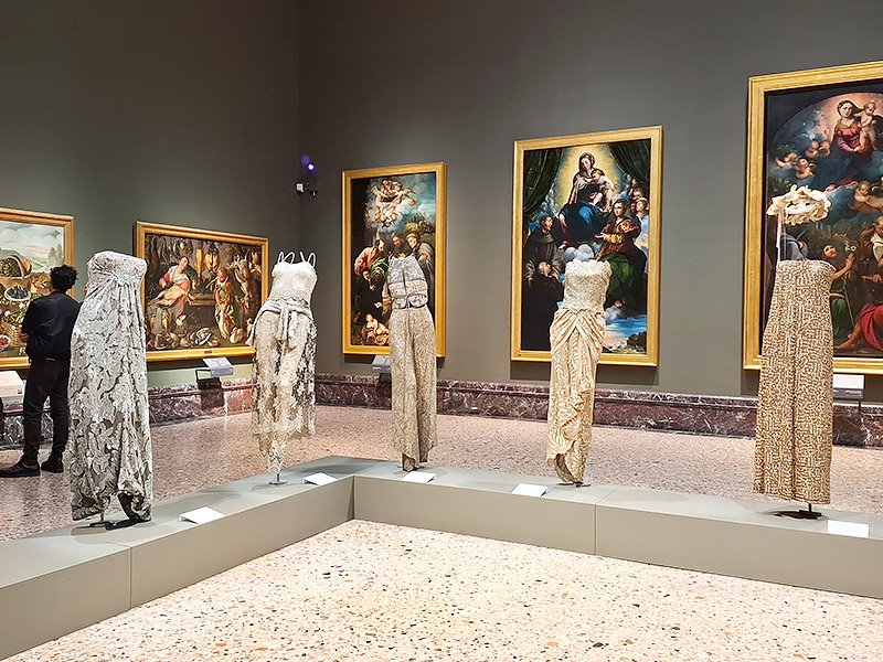 Giorgio Armani fashion creations displayed in a central room of the Pinacoteca di Brera in Milan, with classical paintings in the background.