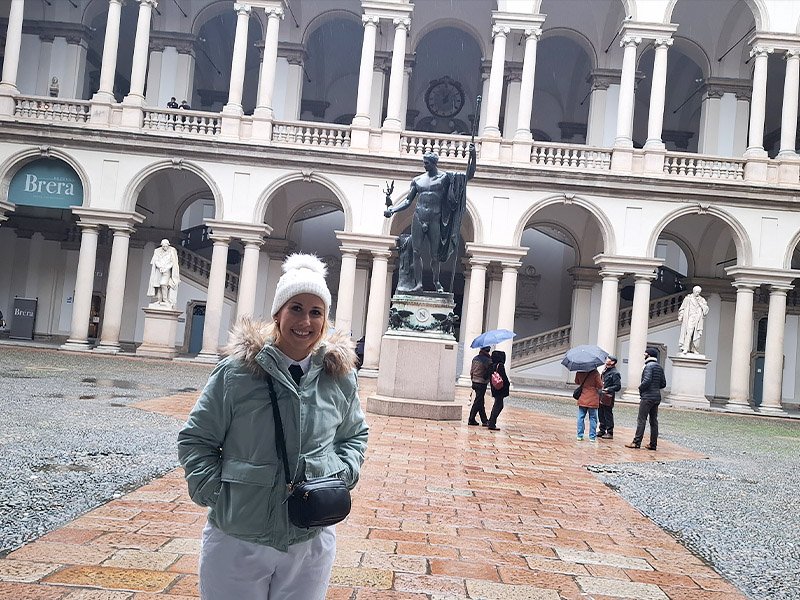 Traveler standing in the courtyard at the entrance of Pinacoteca di Brera in Milan, with the building’s famous arches and central sculpture in the background.